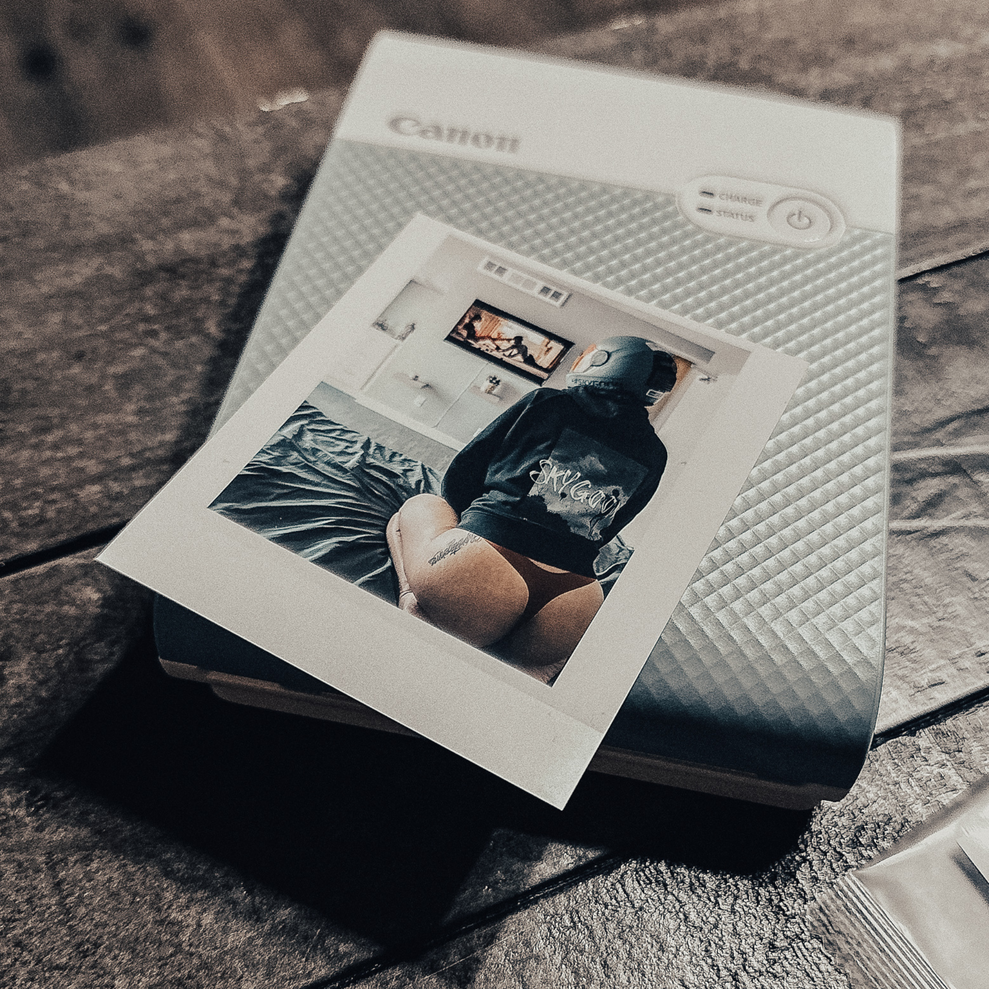 A printed photo of a person in a hoodie and thong sitting on a bed wearing a skydiving helmet, with the photo coming out of a Canon printer, all atop a wooden surface.