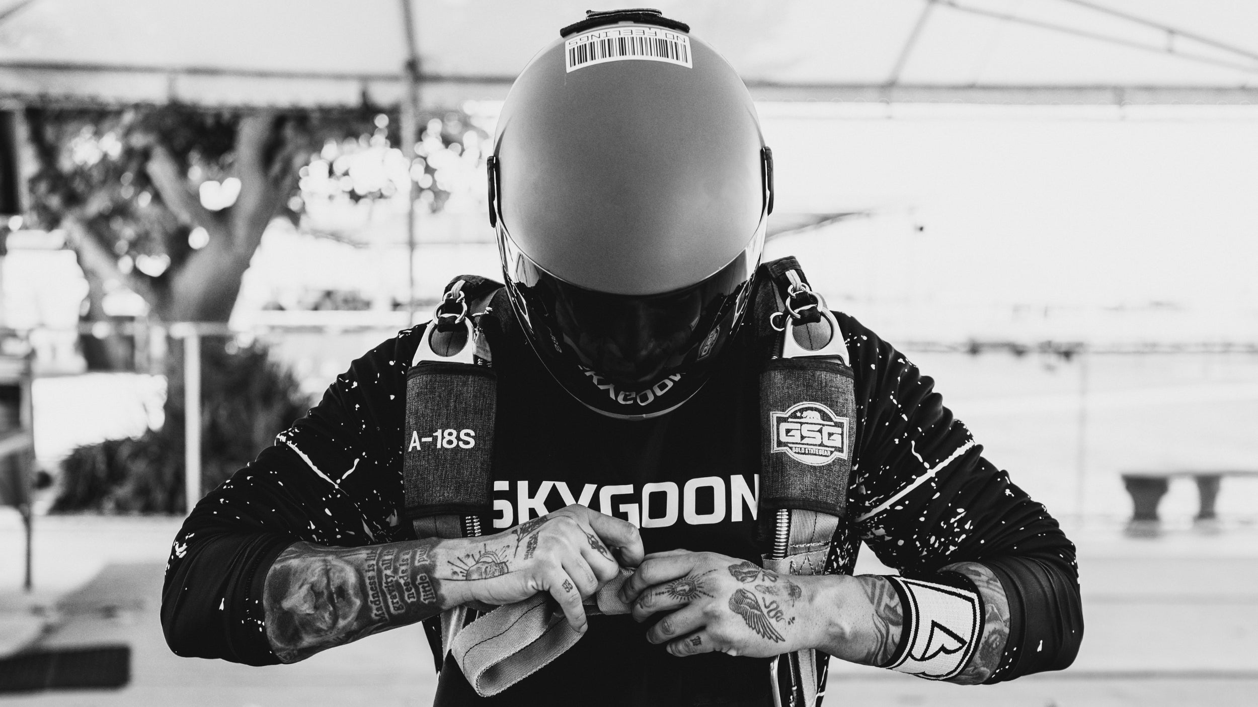 A skydiver checks their gear, focusing on a wrist-mounted device, wearing a full-face helmet and a jumpsuit with visible tattoos on their arms. The image is in black and white.