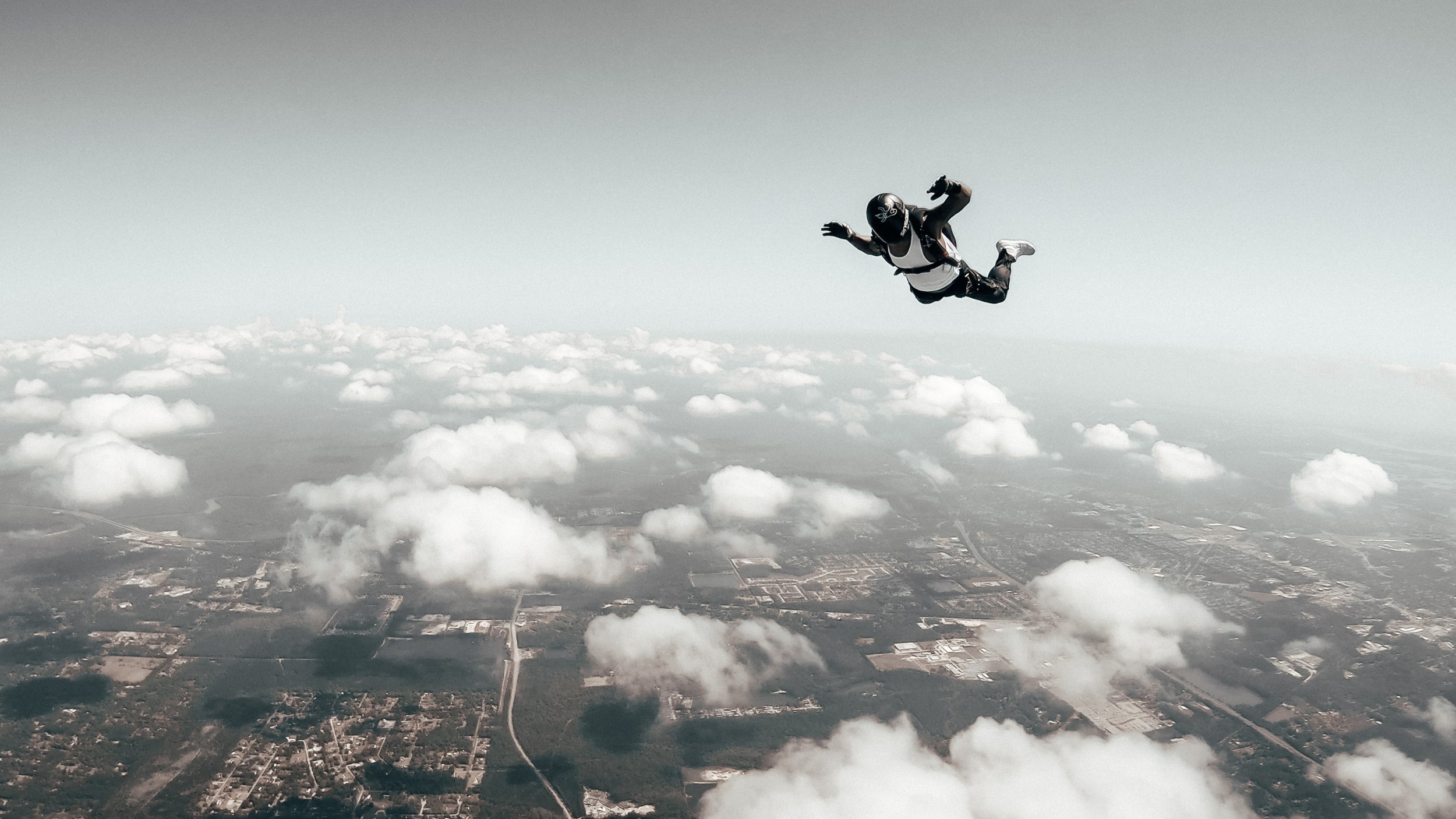 A skydiver freefalls against a backdrop of a vast landscape with scattered clouds, the ground visible far below.