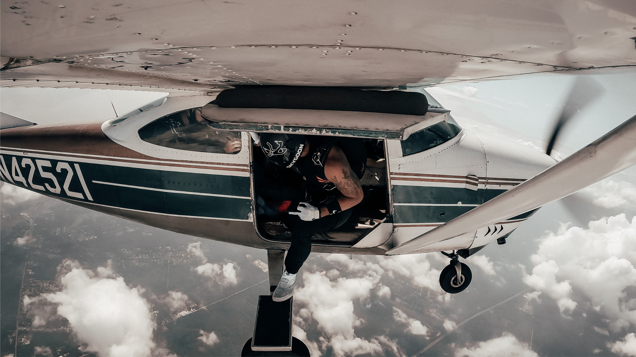 A skydiver in a black jumpsuit prepares to jump from a small airplane, high above a landscape of clouds and land.